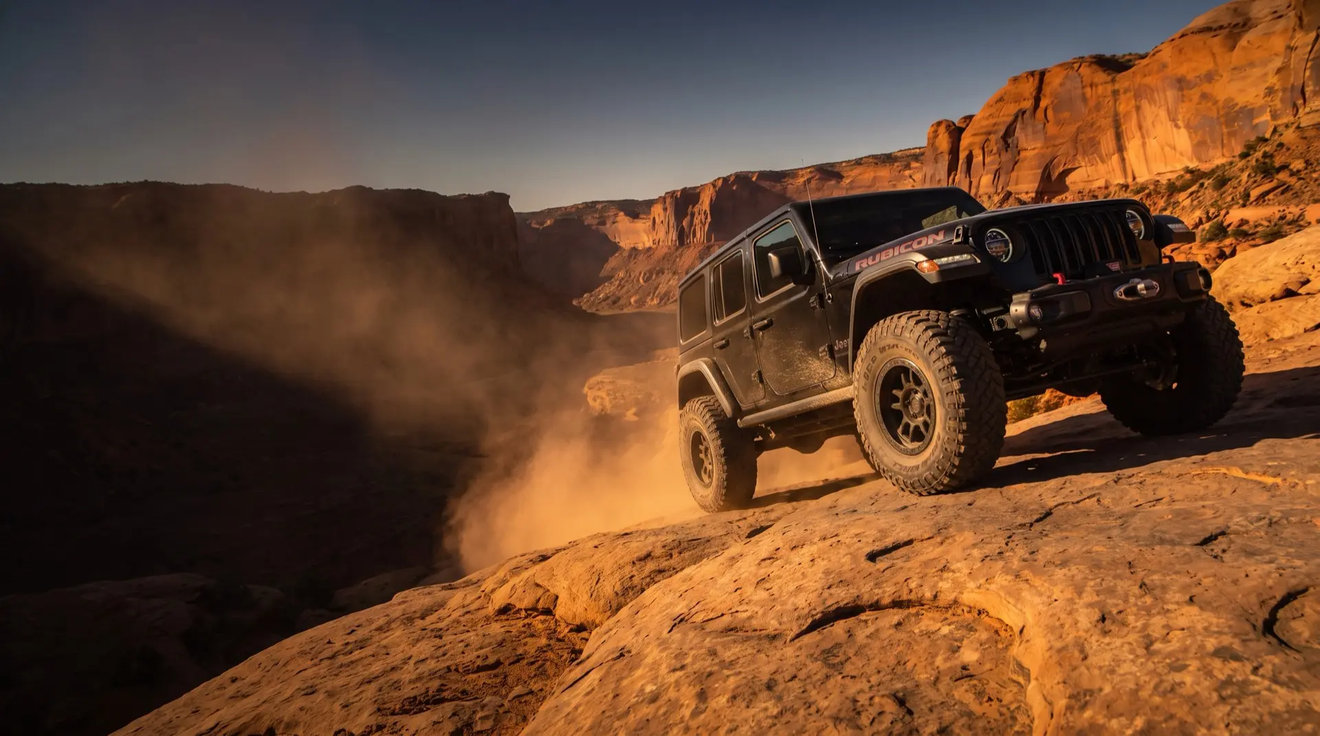 Lifted black Jeep Wrangler Rubicon crawling slickrock in a red-rock canyon at golden hour